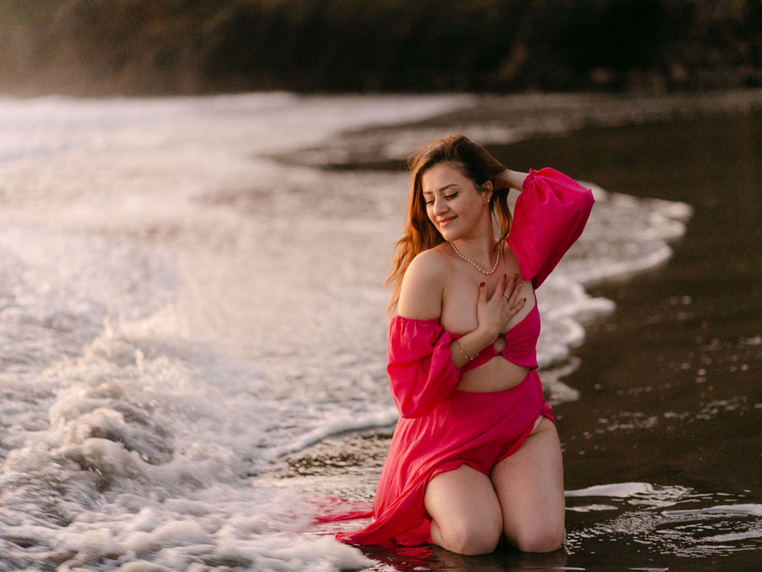A woman in a flowing pink dress kneels in ocean waves on a beach at golden hour.