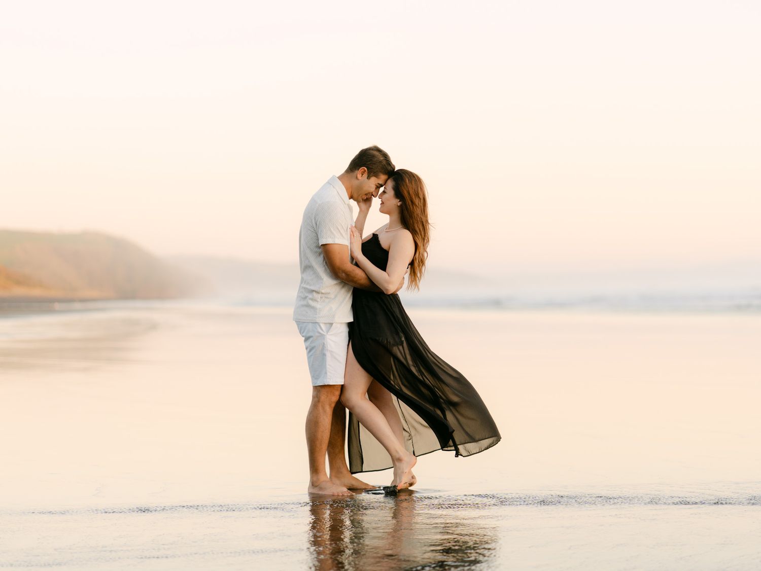 Romantic couple embracing on a serene beach at sunset, waves reflecting warm golden light around their feet.