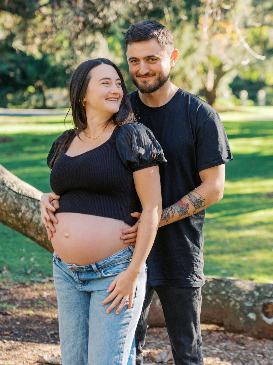Happy pregnant couple embracing outdoors in a park, woman cradling baby bump in black top and jeans.