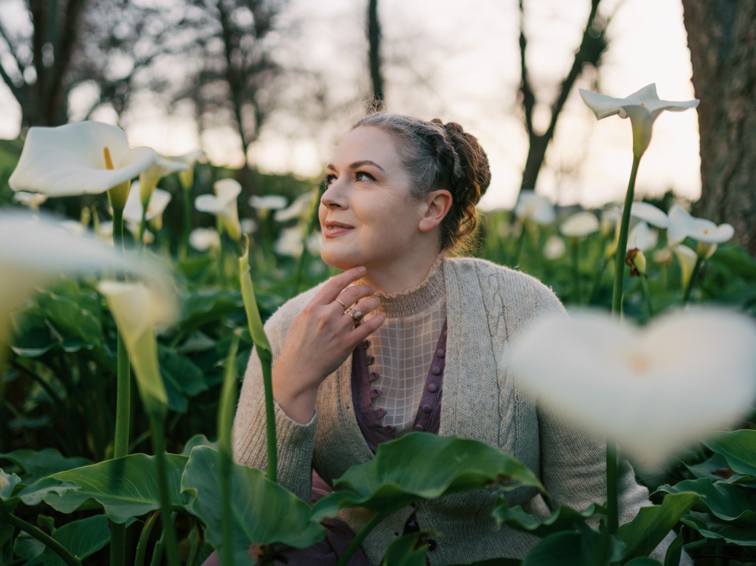 Woman gazing upward among white calla lilies at sunset, surrounded by lush green foliage in a serene garden.