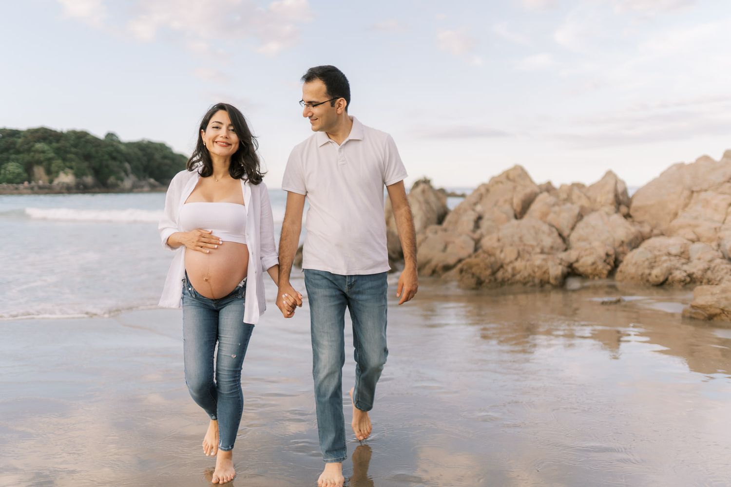 Pregnant woman and man walking hand in hand on a beach, smiling, with rocky cliffs and ocean waves in the background.