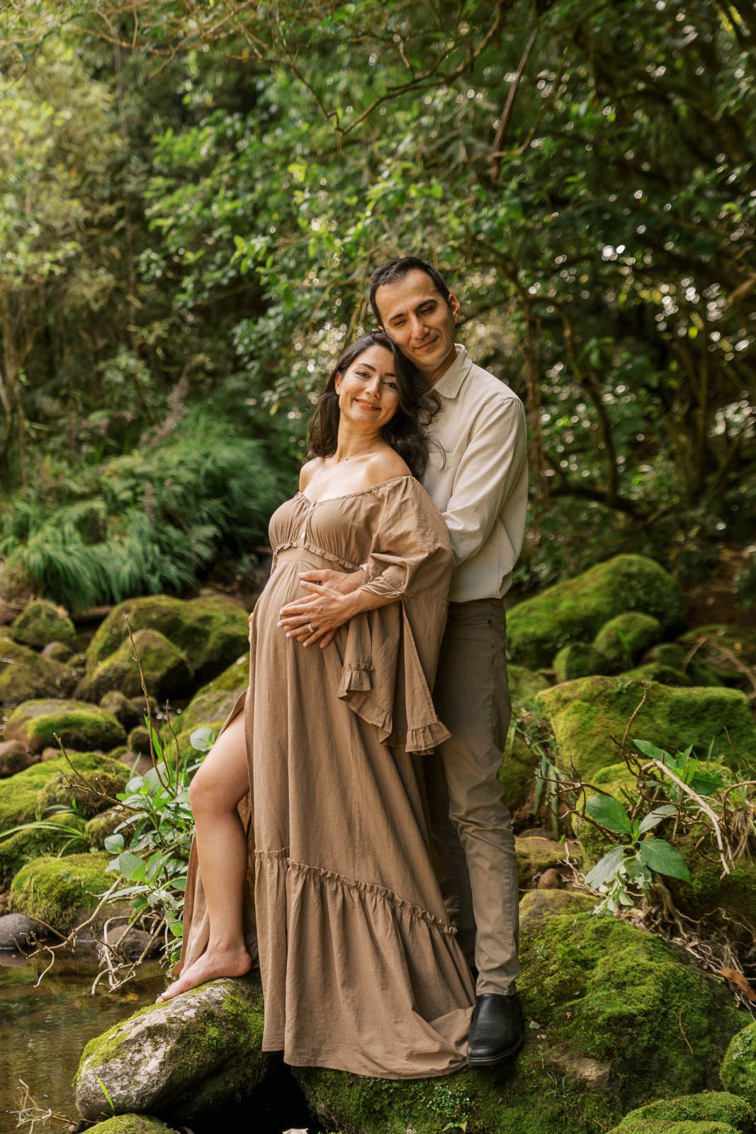 Pregnant couple embracing on mossy rocks in a lush forest, woman in flowing beige dress, man in neutral tones.