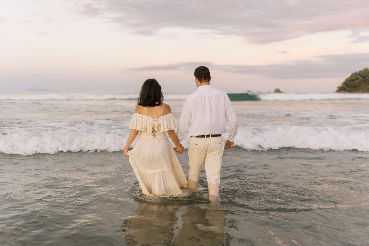 Couple holding hands walking into ocean waves at sunset, woman in white off-shoulder dress, man in white shirt and pants.