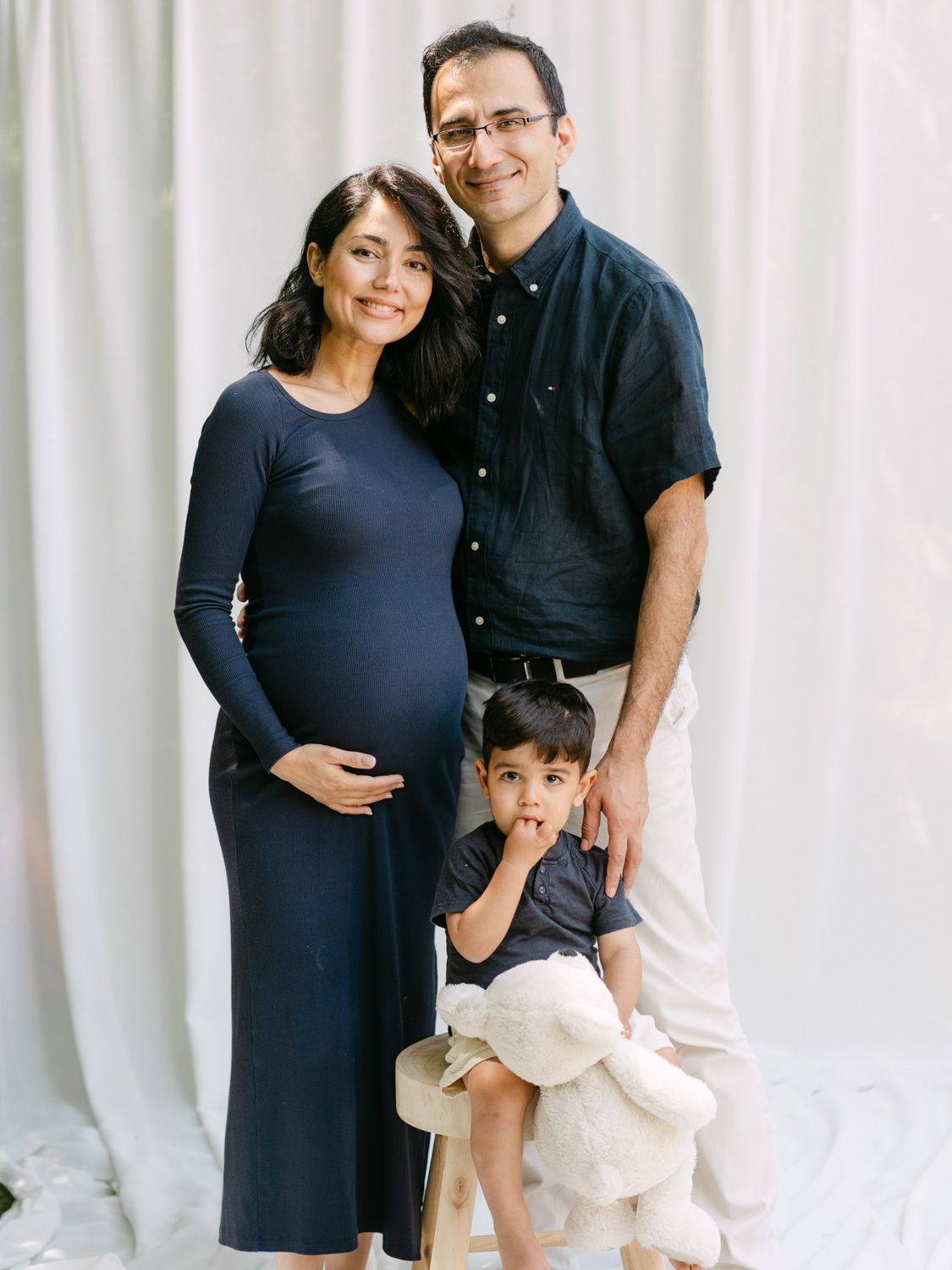 Pregnant woman in navy dress poses with husband and toddler son for a family maternity photo shoot.
