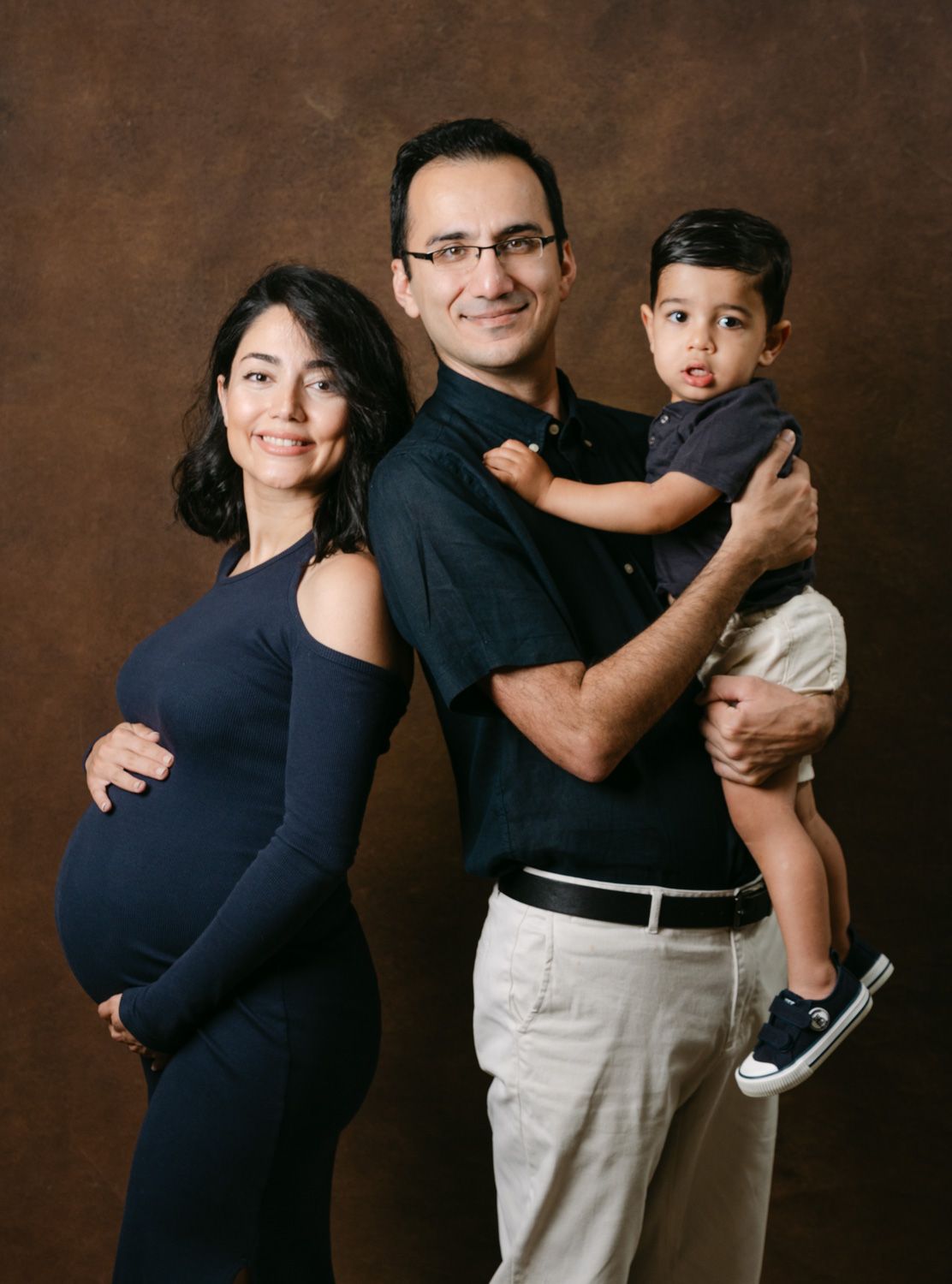 Pregnant woman in navy dress poses with husband holding toddler son in professional family maternity studio portrait.