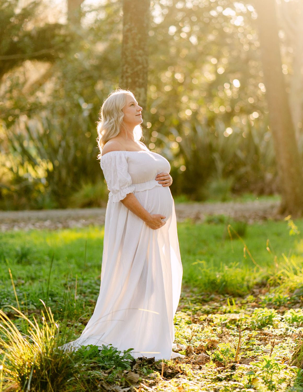 Pregnant woman in white off-shoulder dress standing in sunlit garden, cradling baby bump, golden hour photography.