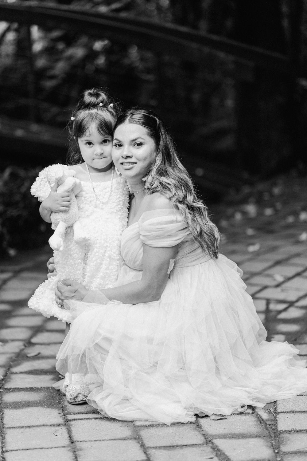 Black and white photo of a mother in a white gown crouching beside her young daughter in a white dress outdoors.