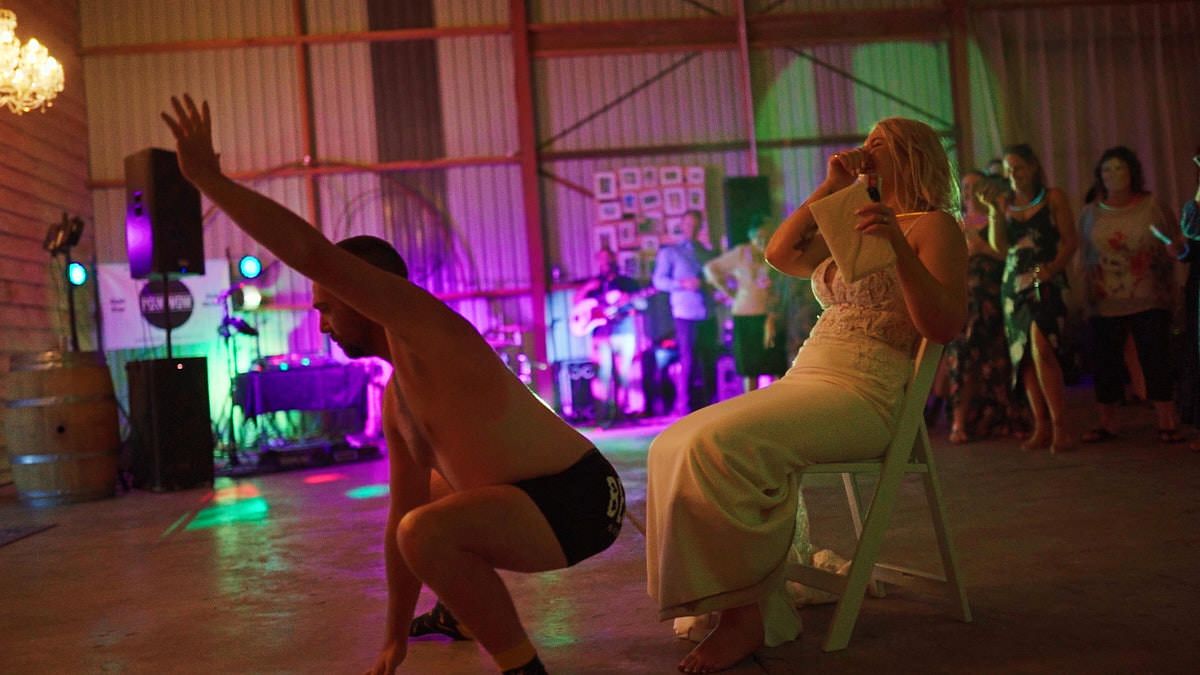 Shirtless groom performing a chair dance for bride in white dress at rustic wedding reception with live band.