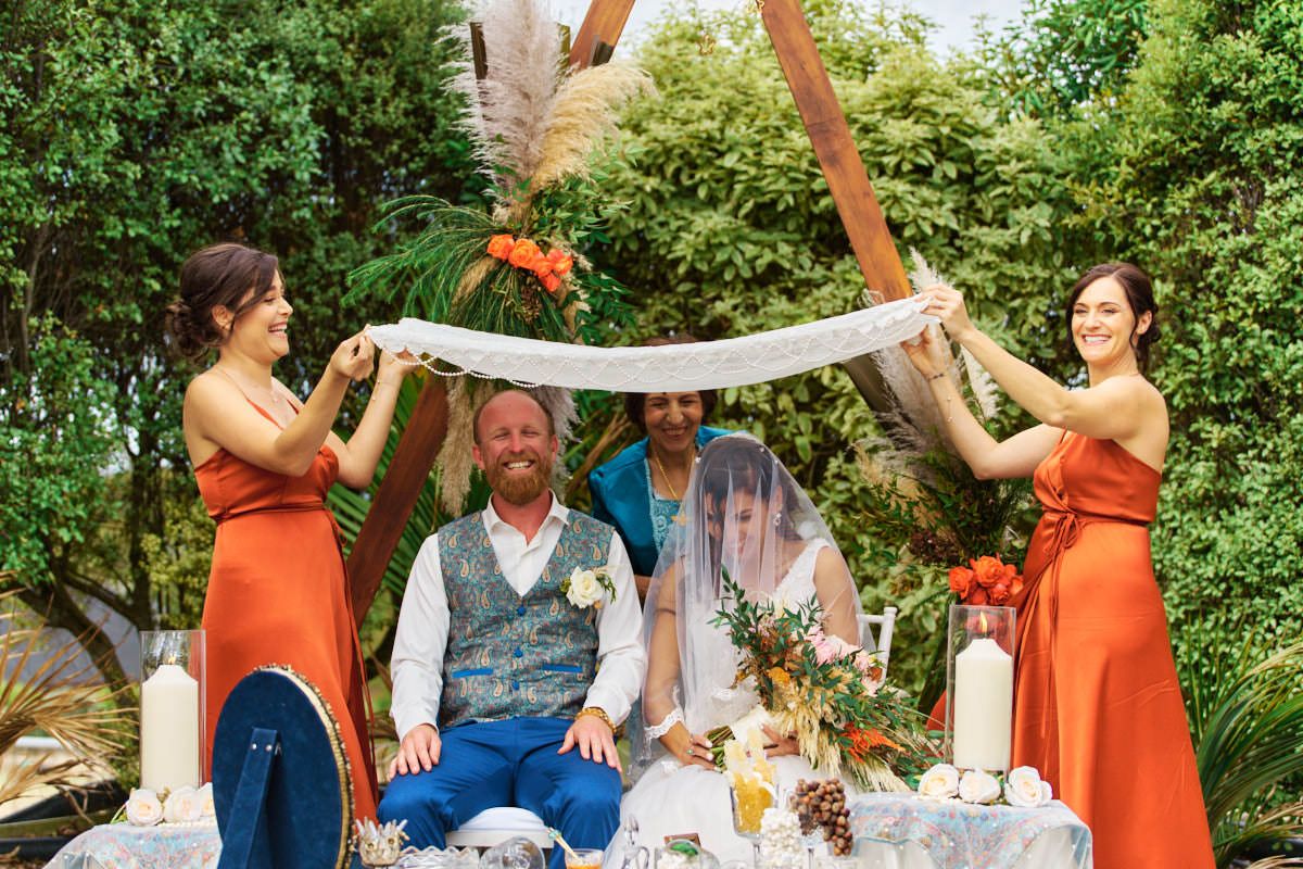 Bridesmaids in orange dresses hold fabric over groom seated at outdoor wedding ceremony table with floral decor.