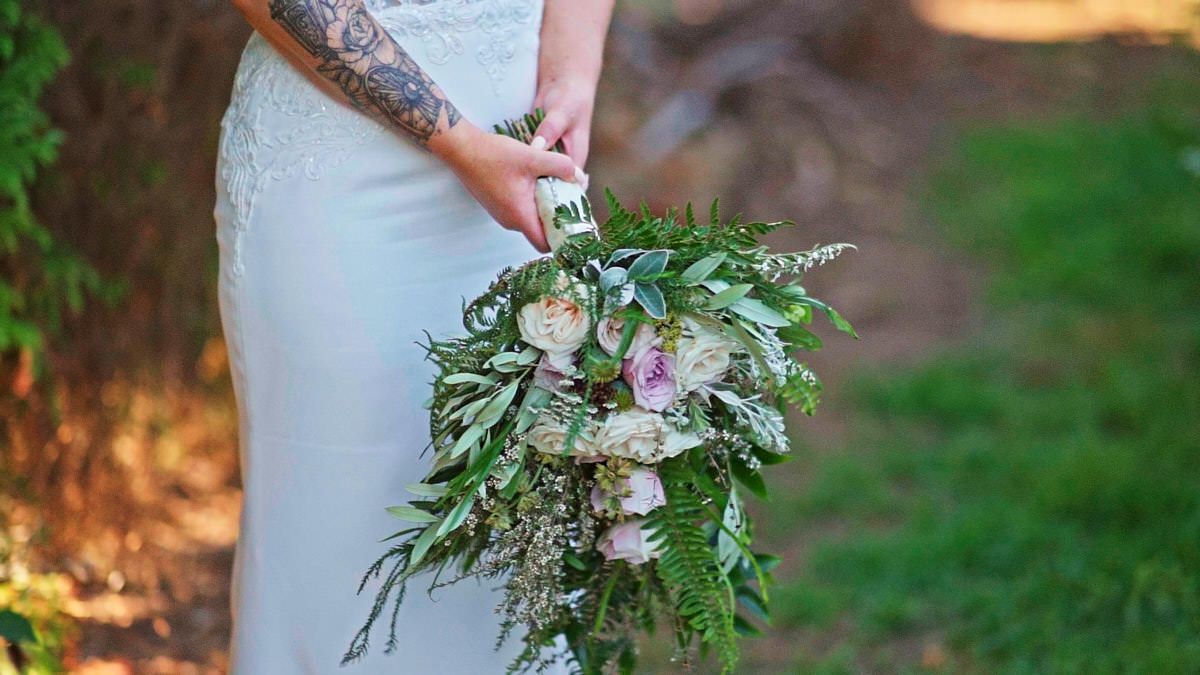 Bride holding a lush cascading wedding bouquet with white flowers and greenery in natural outdoor setting.