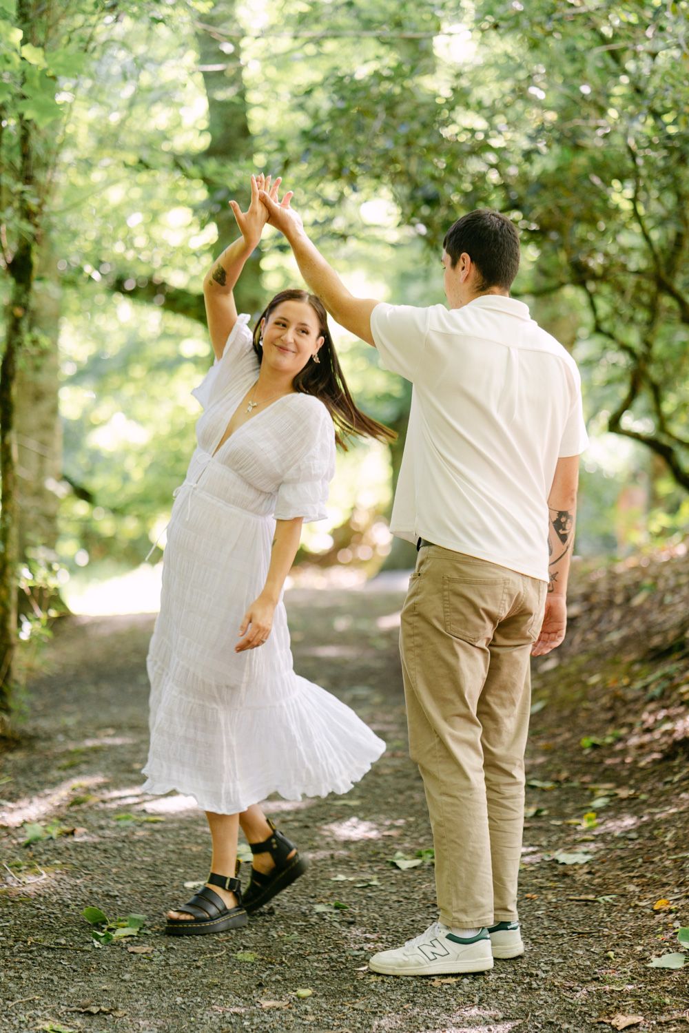 Couple dancing on a forest path, woman in white dress being twirled by man in beige pants amid lush green trees.