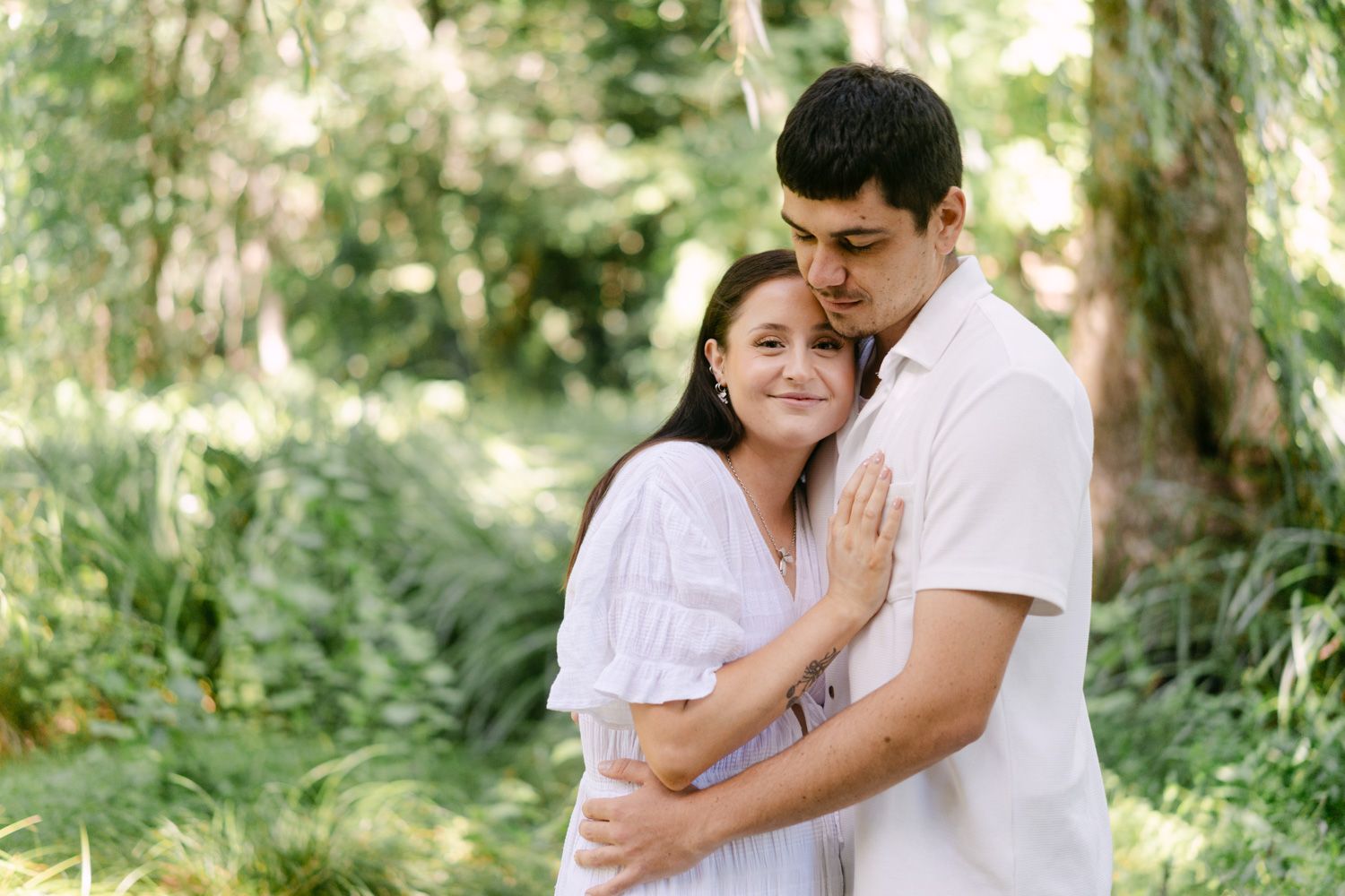 A couple embraces tenderly in a lush green garden, the man kissing the woman's head as she smiles softly.