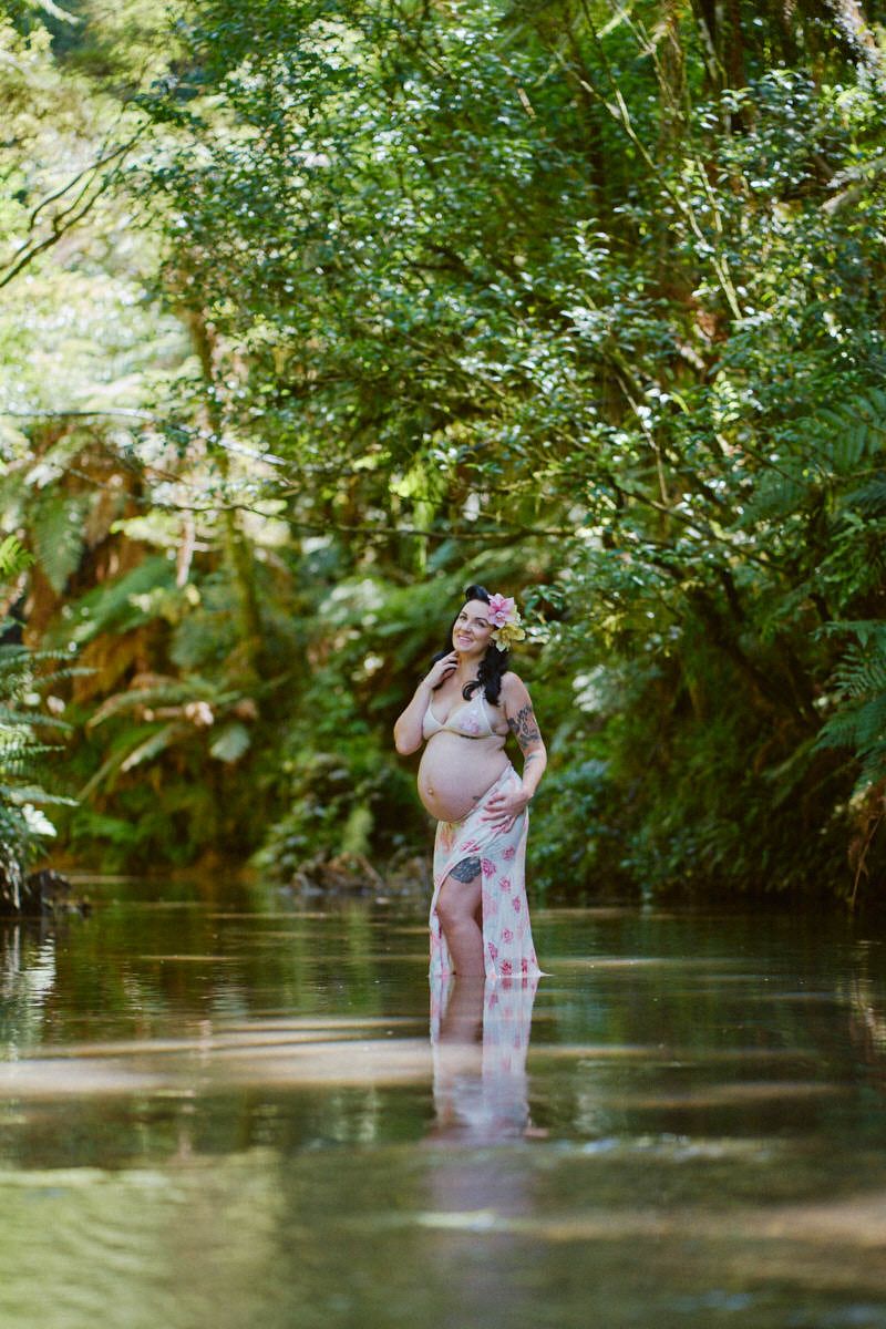 Pregnant woman standing in a forest stream wearing a floral wrap dress and flower crown, cradling her baby bump.