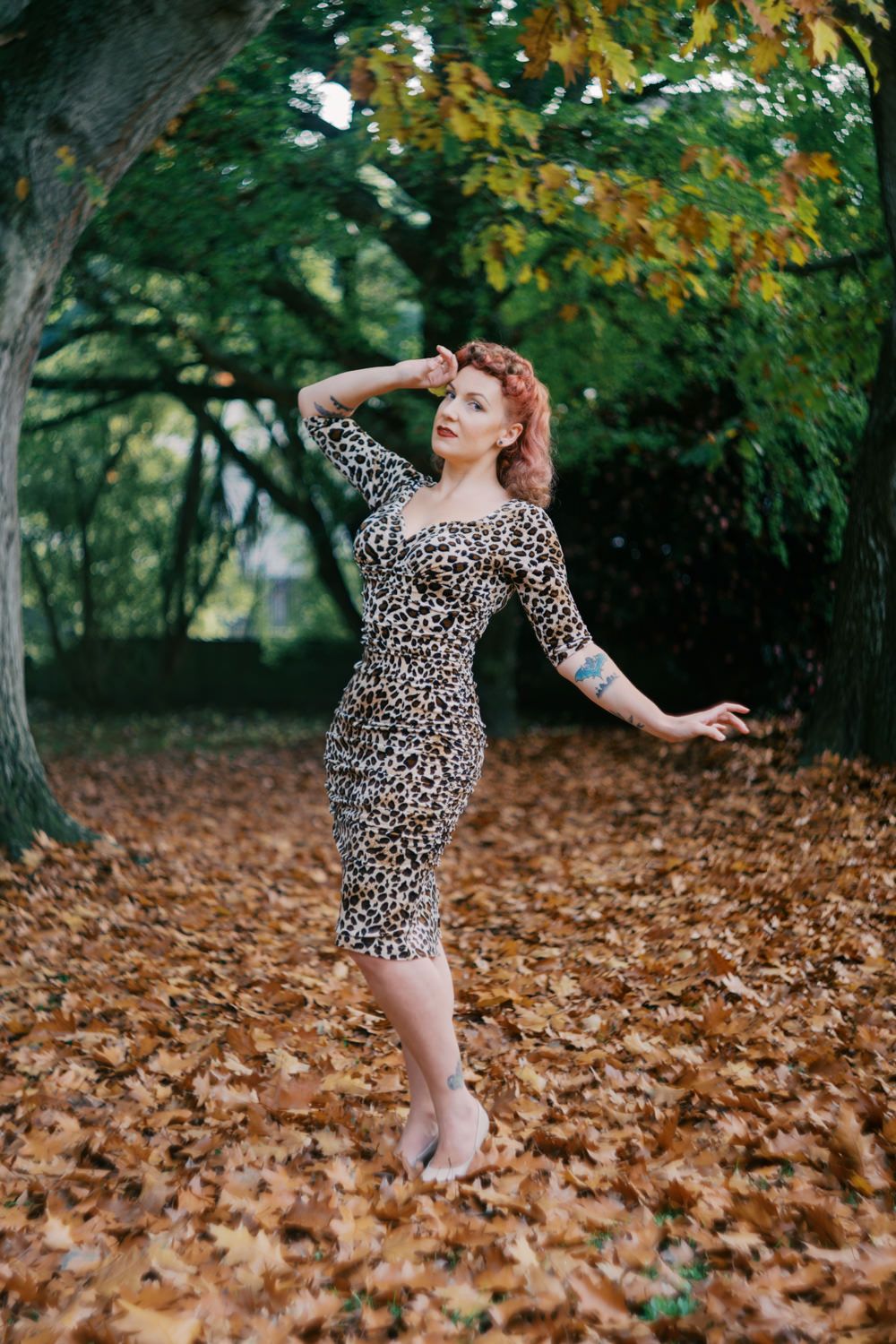 Woman in leopard print dress poses among autumn leaves in a park, with trees displaying fall foliage behind her.