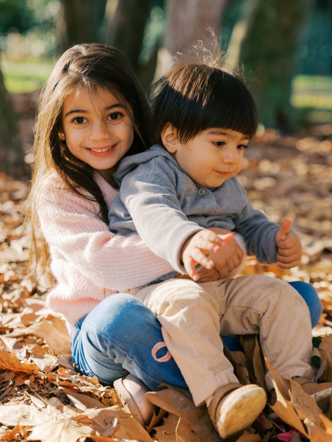 Two young siblings sitting on autumn leaves, older girl hugging younger toddler boy, smiling outdoors in a park.