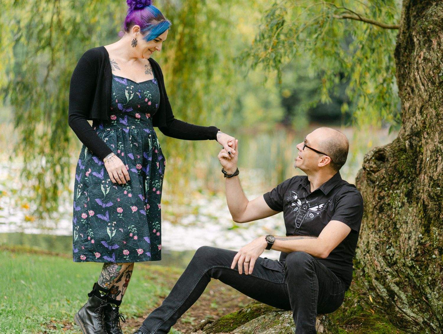 A couple shares a romantic moment under a willow tree, woman in floral dress holding hands with seated man in black.