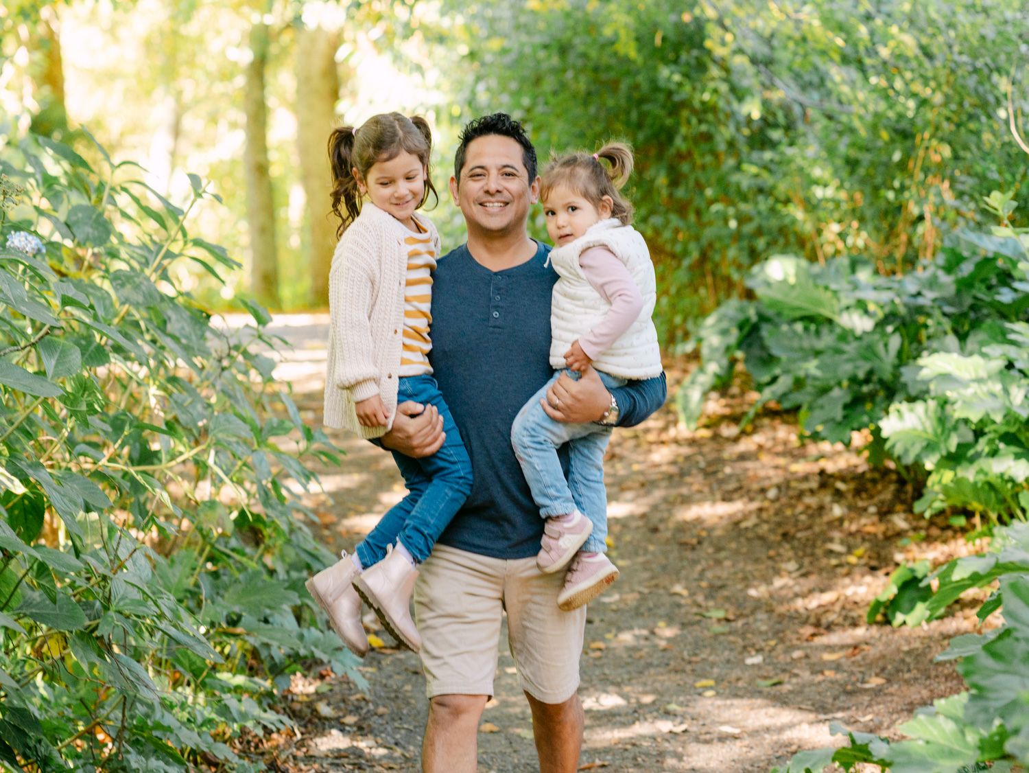 A smiling man holds two young girls on a sunny tree-lined path surrounded by lush green foliage.