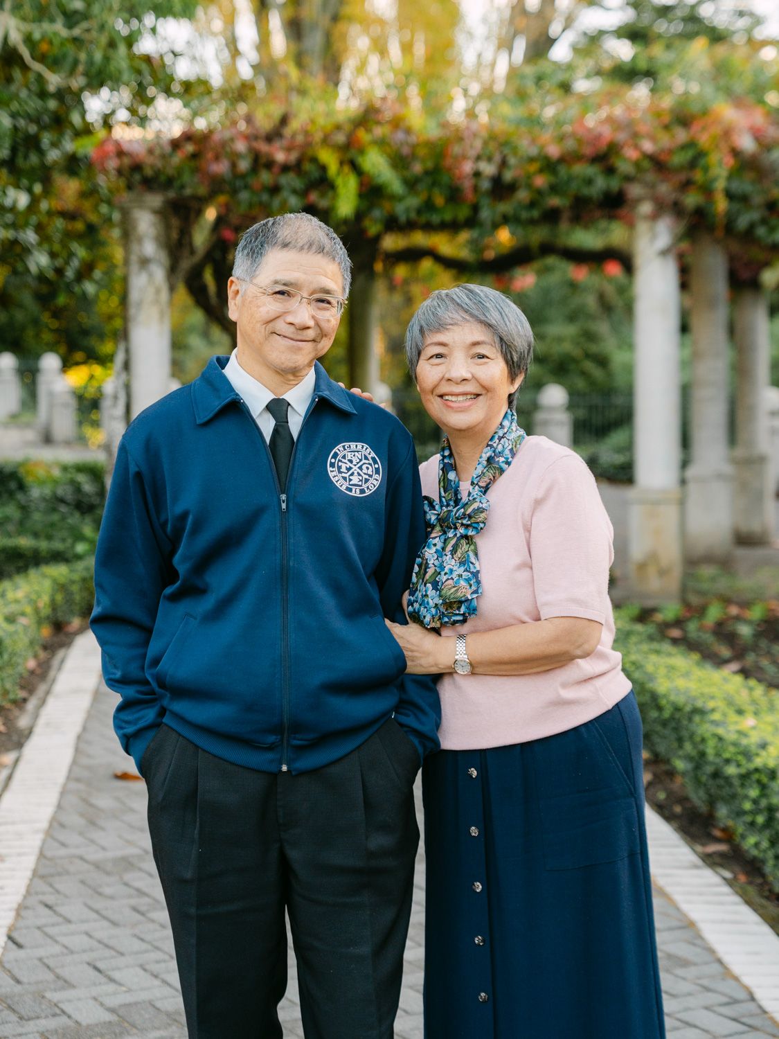 Elderly Asian couple smiling and posing together on a garden pathway surrounded by lush greenery and flowers.
