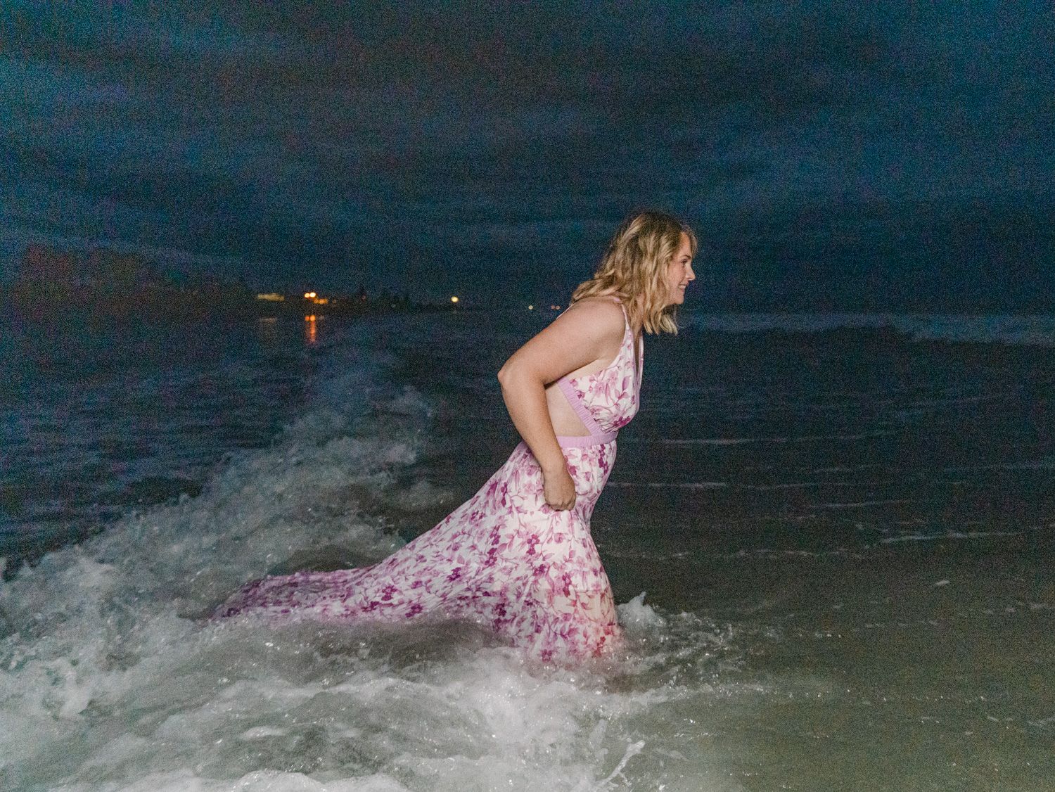 Woman in floral pink dress wading through ocean waves at night on a dark beach.