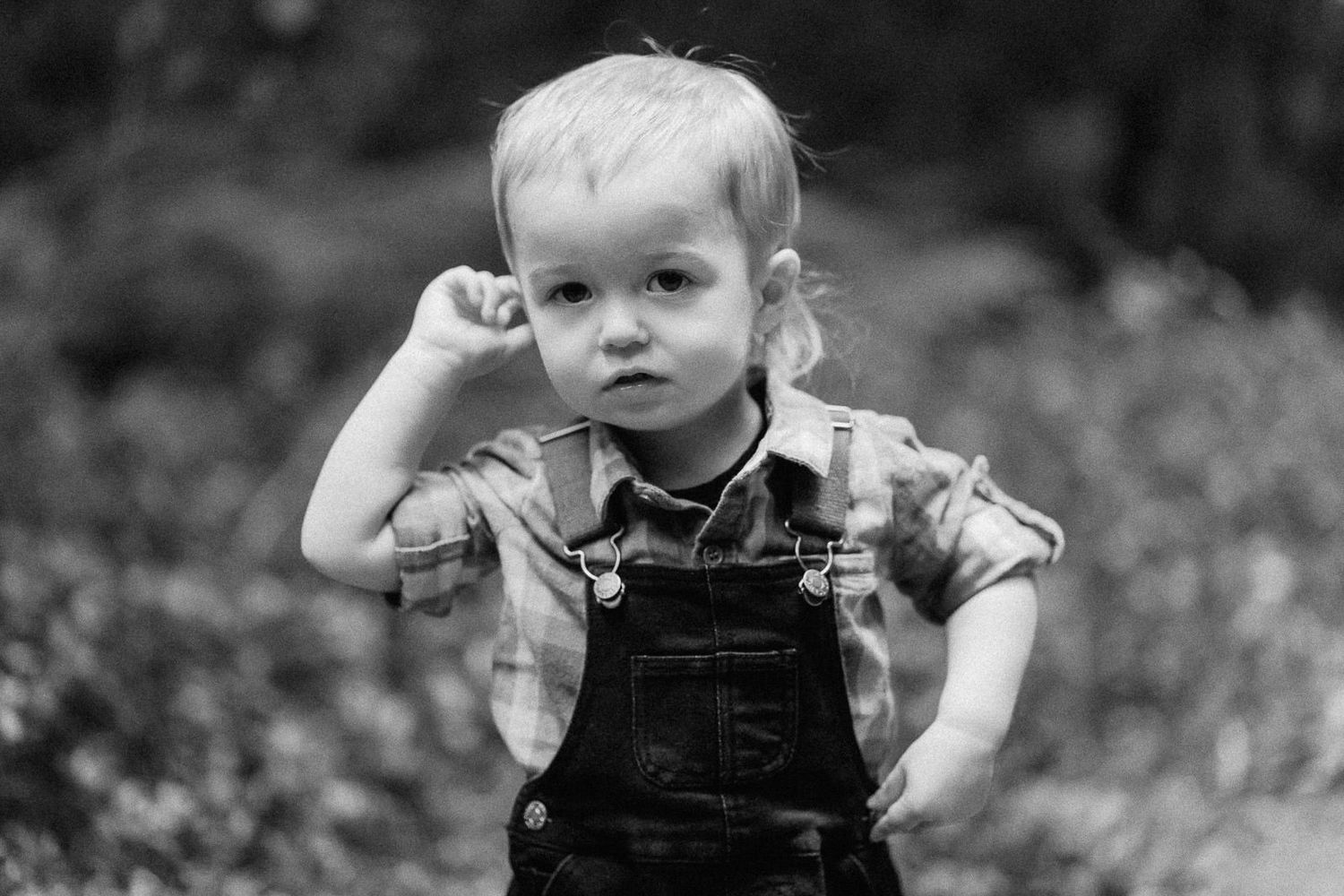 Black and white portrait of a toddler in overalls and plaid shirt posing outdoors.