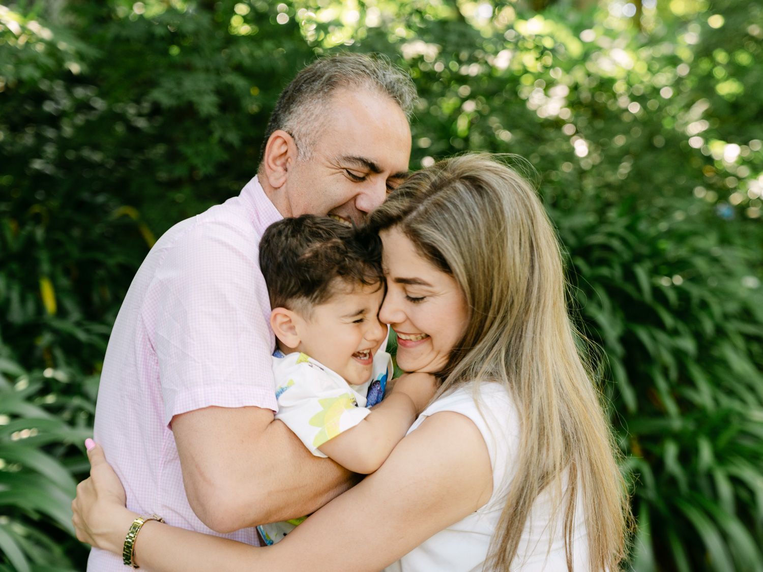 A loving family of three sharing a warm embrace outdoors, with parents hugging their young toddler among lush green foliage.