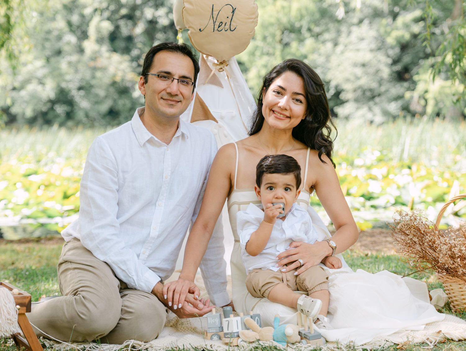 Family of three posing outdoors on a picnic blanket, holding a heart-shaped balloon that reads Neil.