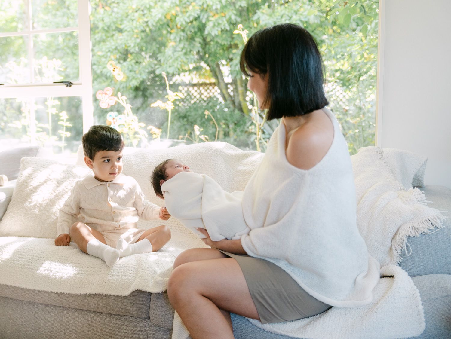 A mother sits on a white sofa holding a newborn while her toddler sits beside her in a bright living room.