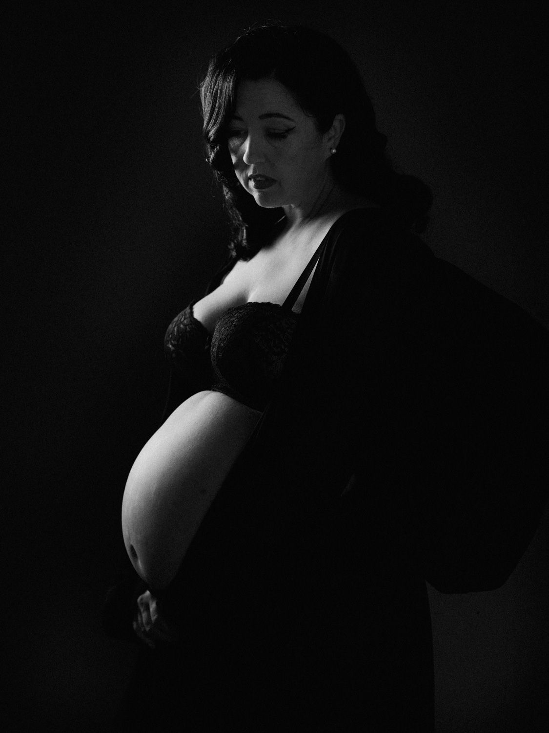 Black and white maternity portrait of a pregnant woman in dark lingerie cradling her baby bump against a dark background.