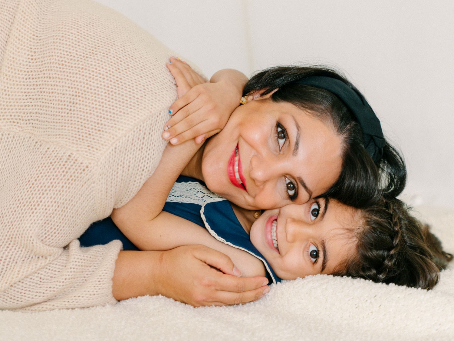 A mother and young daughter share a warm embrace, smiling together while lying on a soft white blanket.