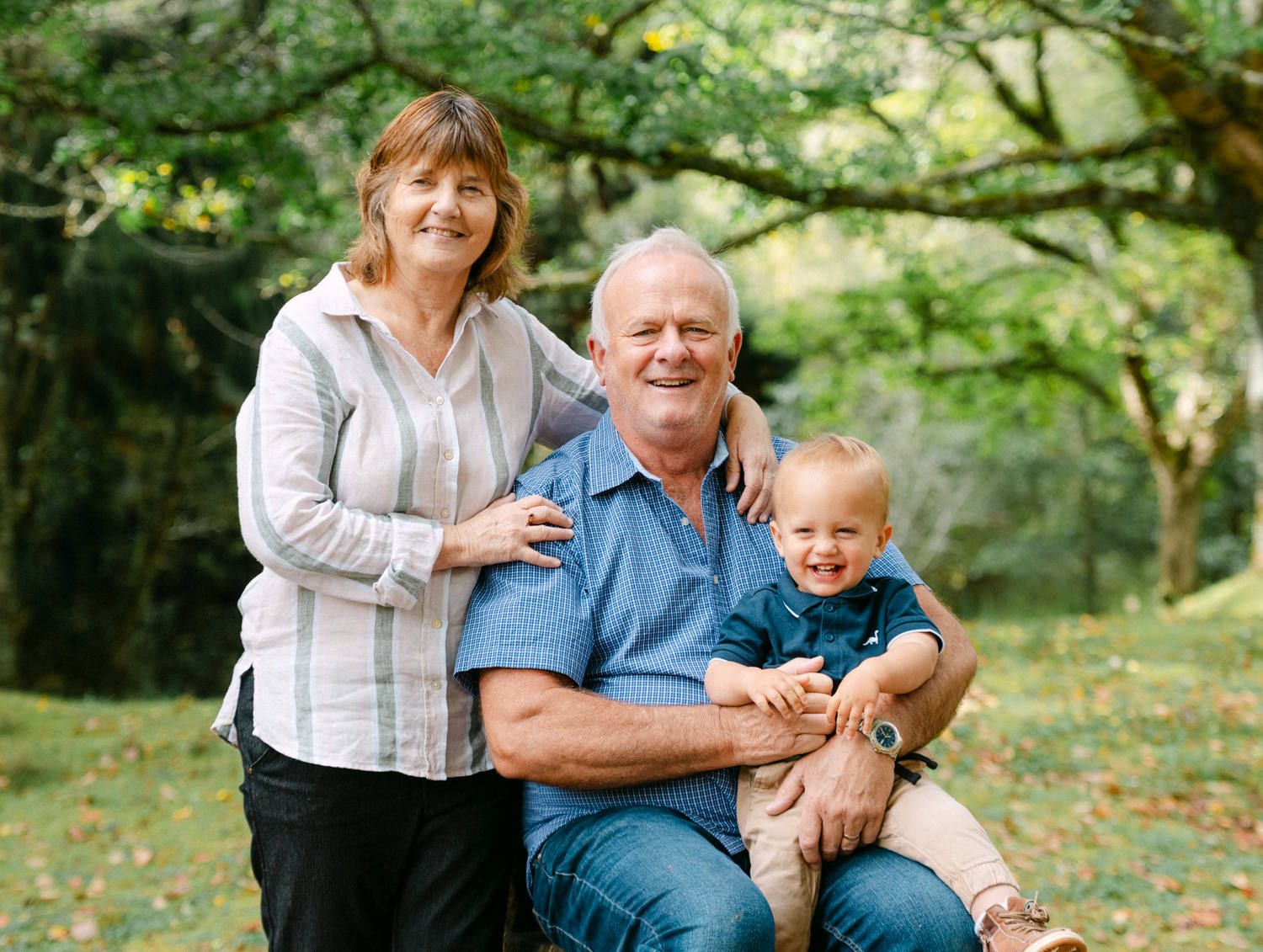 Grandparents smiling outdoors with toddler grandson, sitting under green trees in a lush park setting.