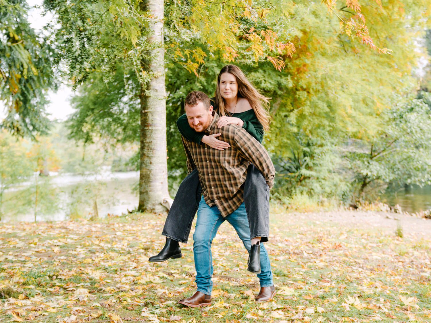 Couple enjoying a piggyback ride in an autumn park with colorful fallen leaves and trees by a lake.