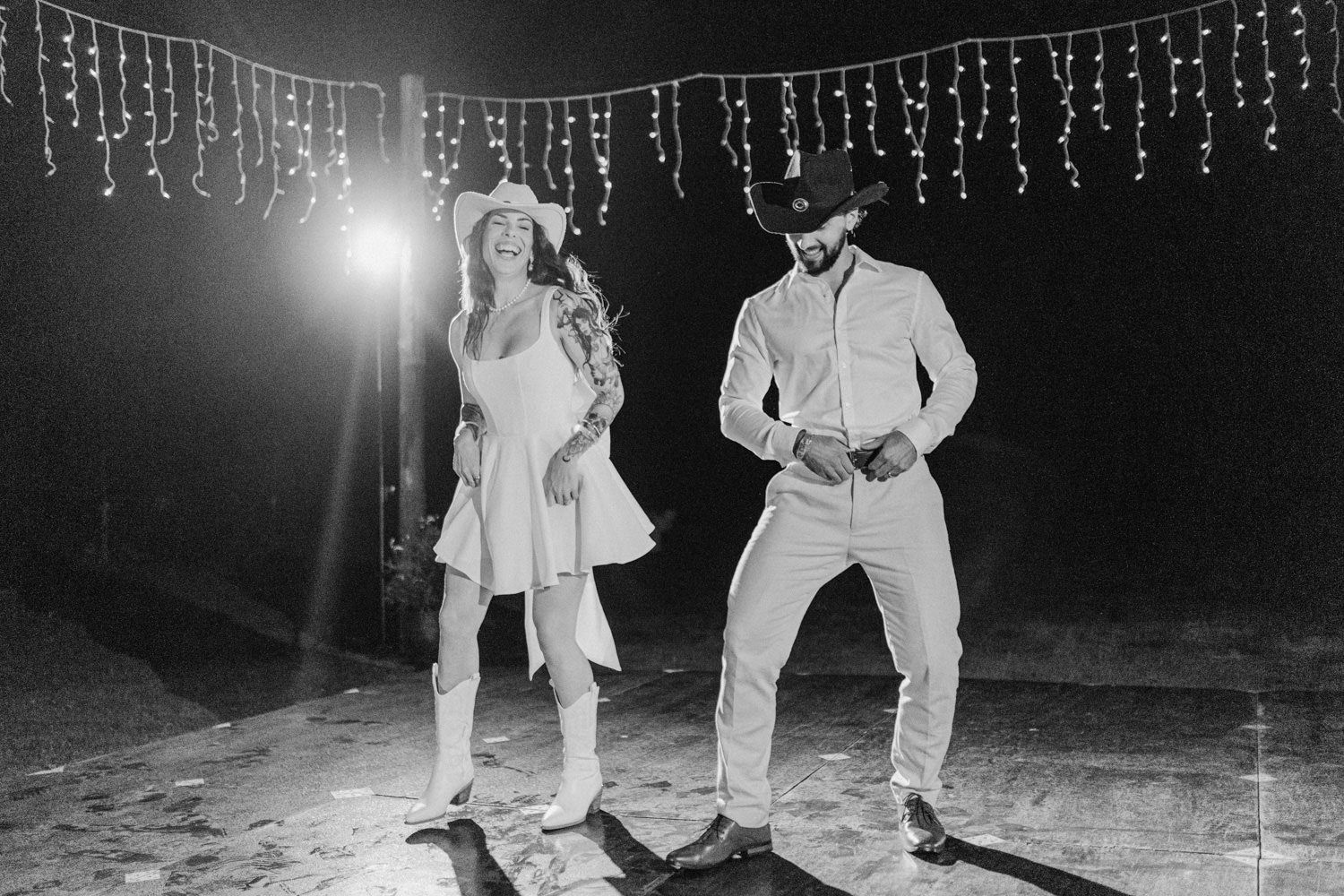 Black and white photo of a couple dancing outdoors at night under string lights, wearing cowboy hats.