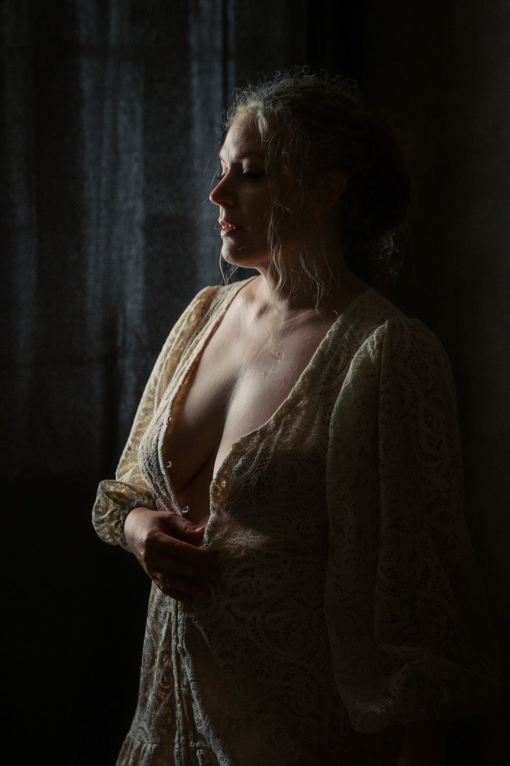 Elegant woman in sheer lace robe standing in dramatic moody window light.