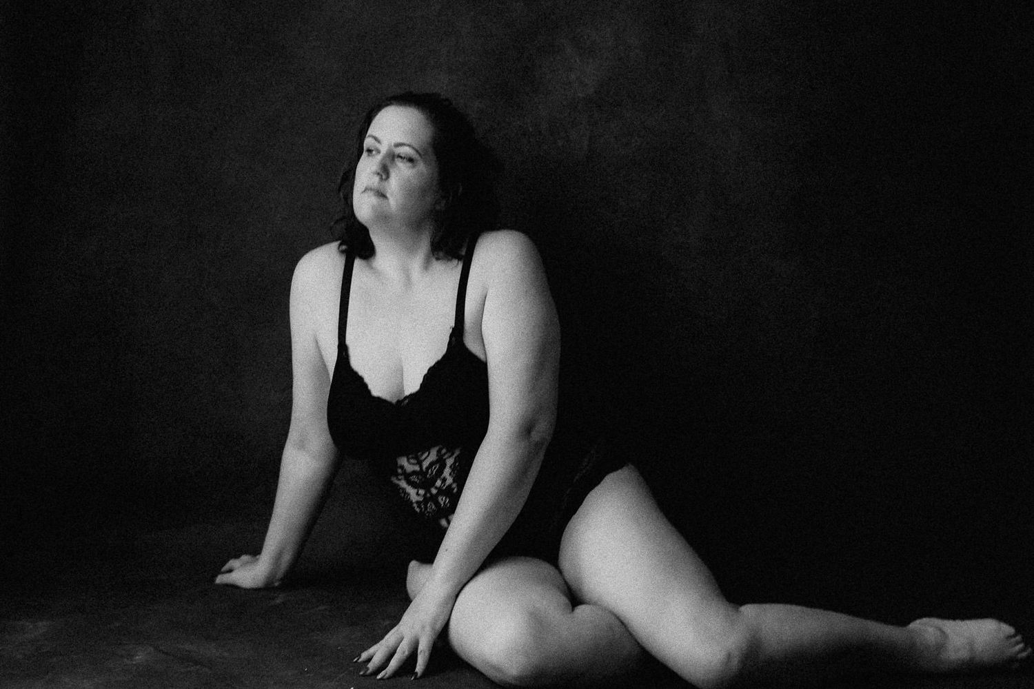 Black and white portrait of a plus-size woman in black bodysuit sitting gracefully on studio floor.