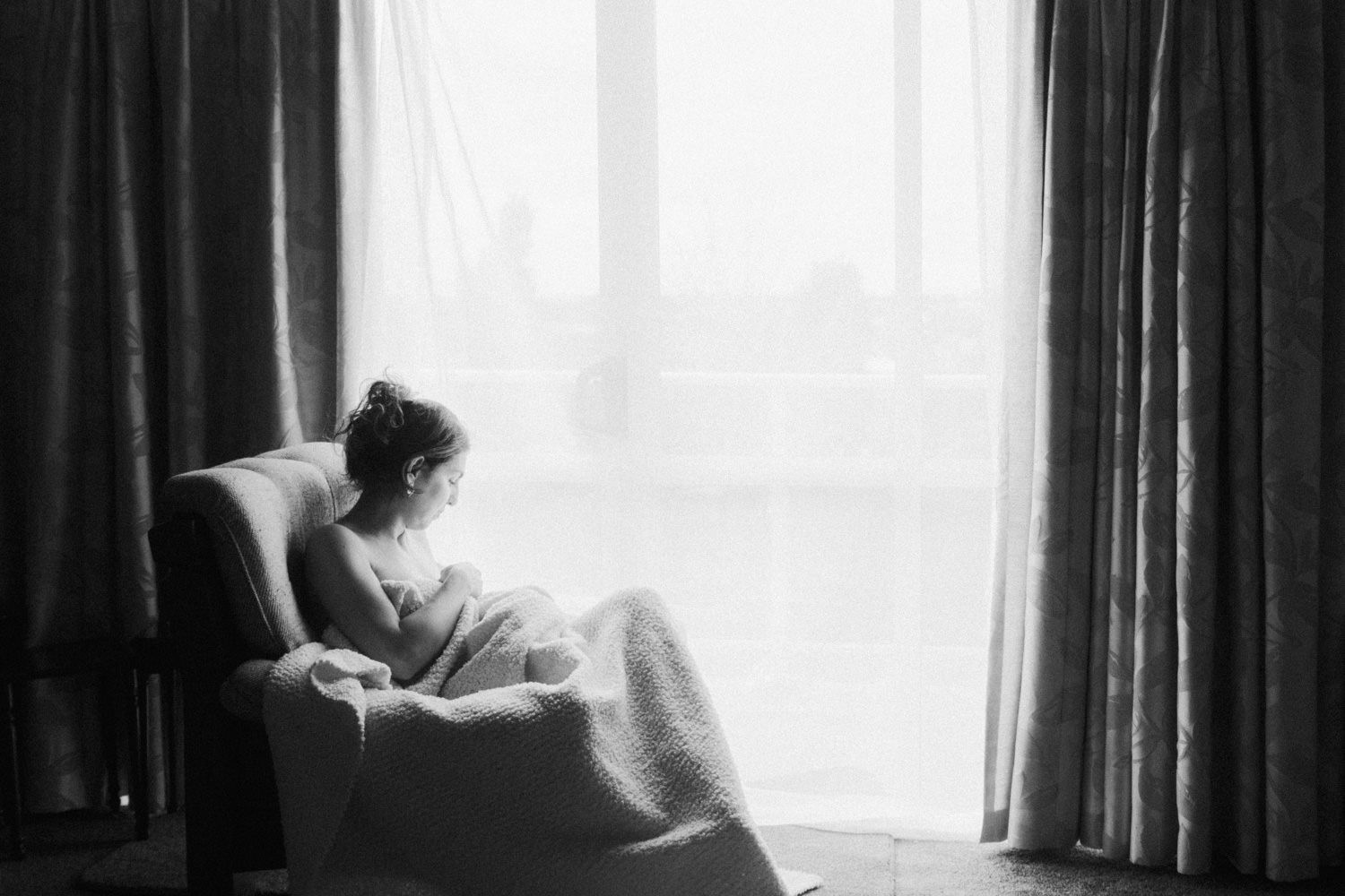 Black and white photo of a woman sitting alone in an armchair by a curtained window, gazing outside contemplatively.