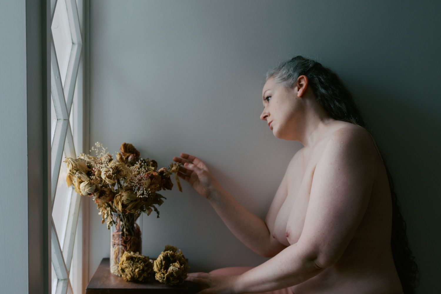 A woman with blue hair arranges dried flowers in a vase by a window in soft natural light.