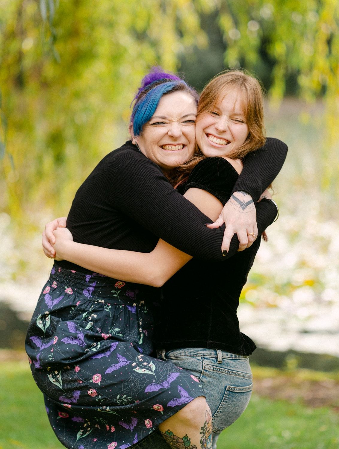 Two women hugging and smiling outdoors, one with blue and purple hair, wearing black tops, in a lush green park setting.