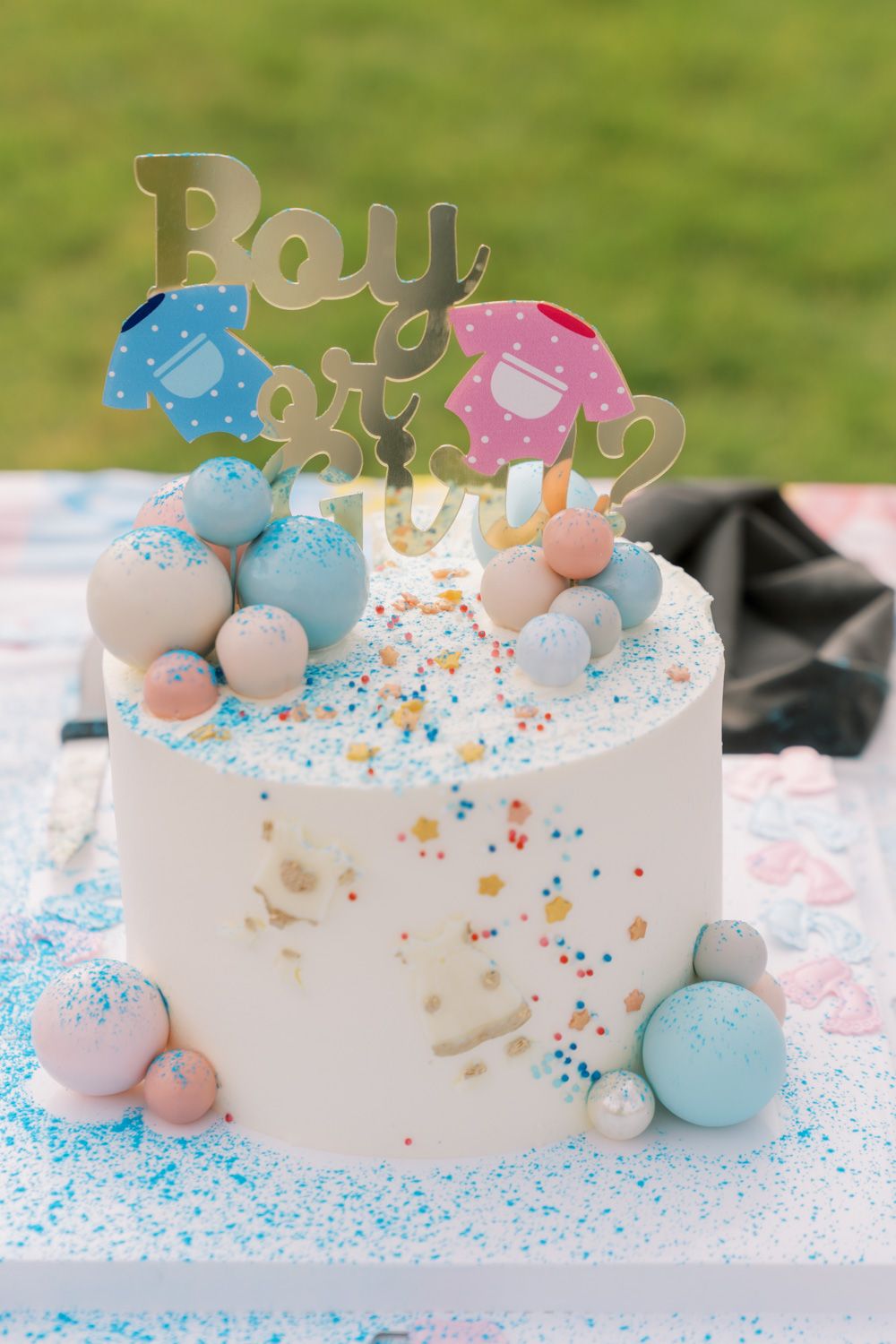Gender reveal cake with blue and pink decorations, macarons, and a Boy or Girl topper on a white frosted cake.