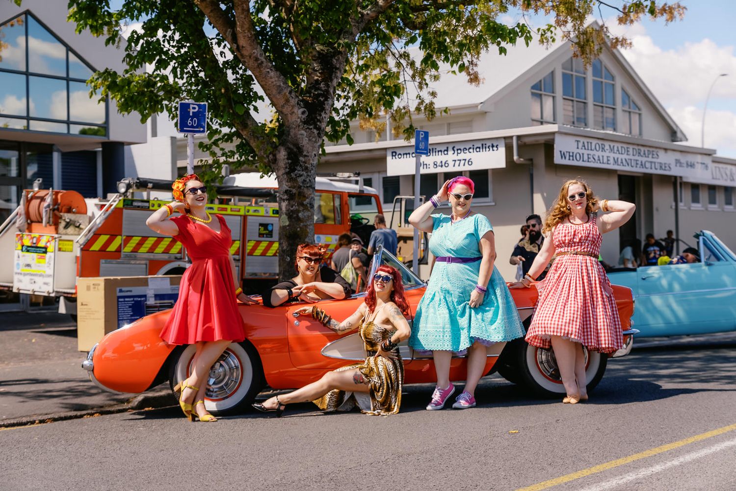 Women in colorful vintage 1950s costumes pose beside a classic red car during a sunny outdoor parade event.