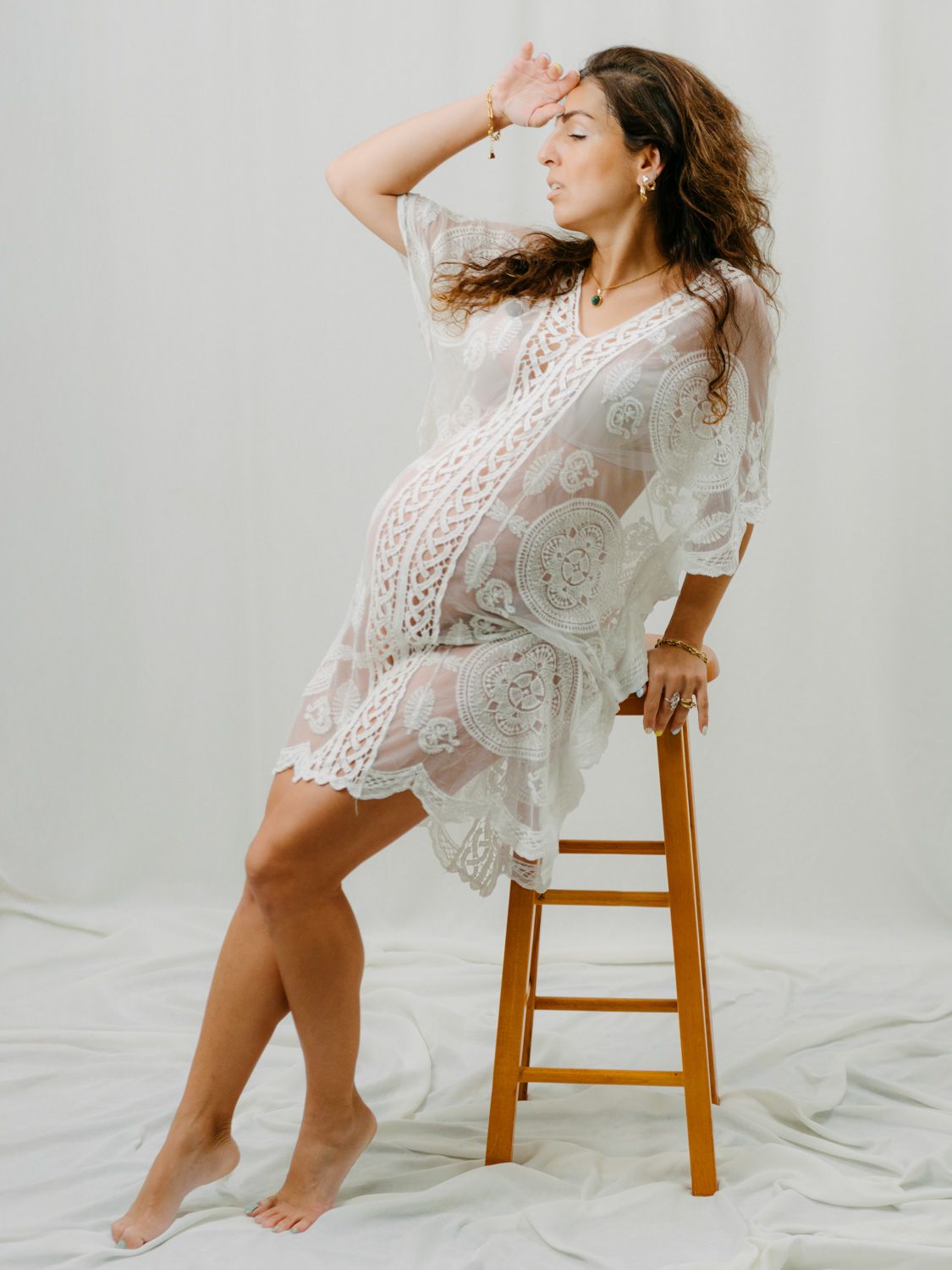 Pregnant woman in white lace dress poses on wooden stool in studio maternity photoshoot.
