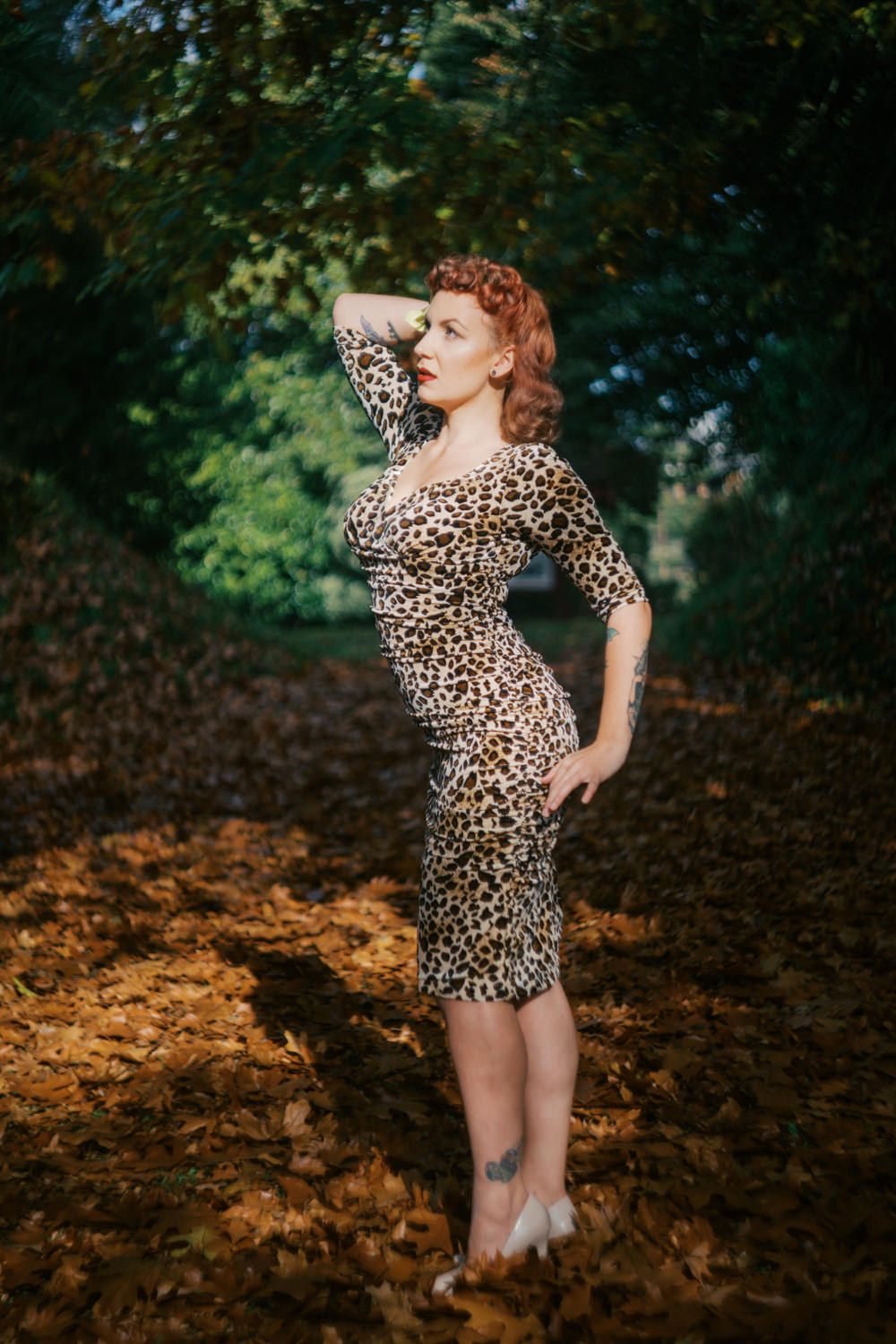 Woman in leopard print dress posing among autumn leaves in a sunlit forest setting.