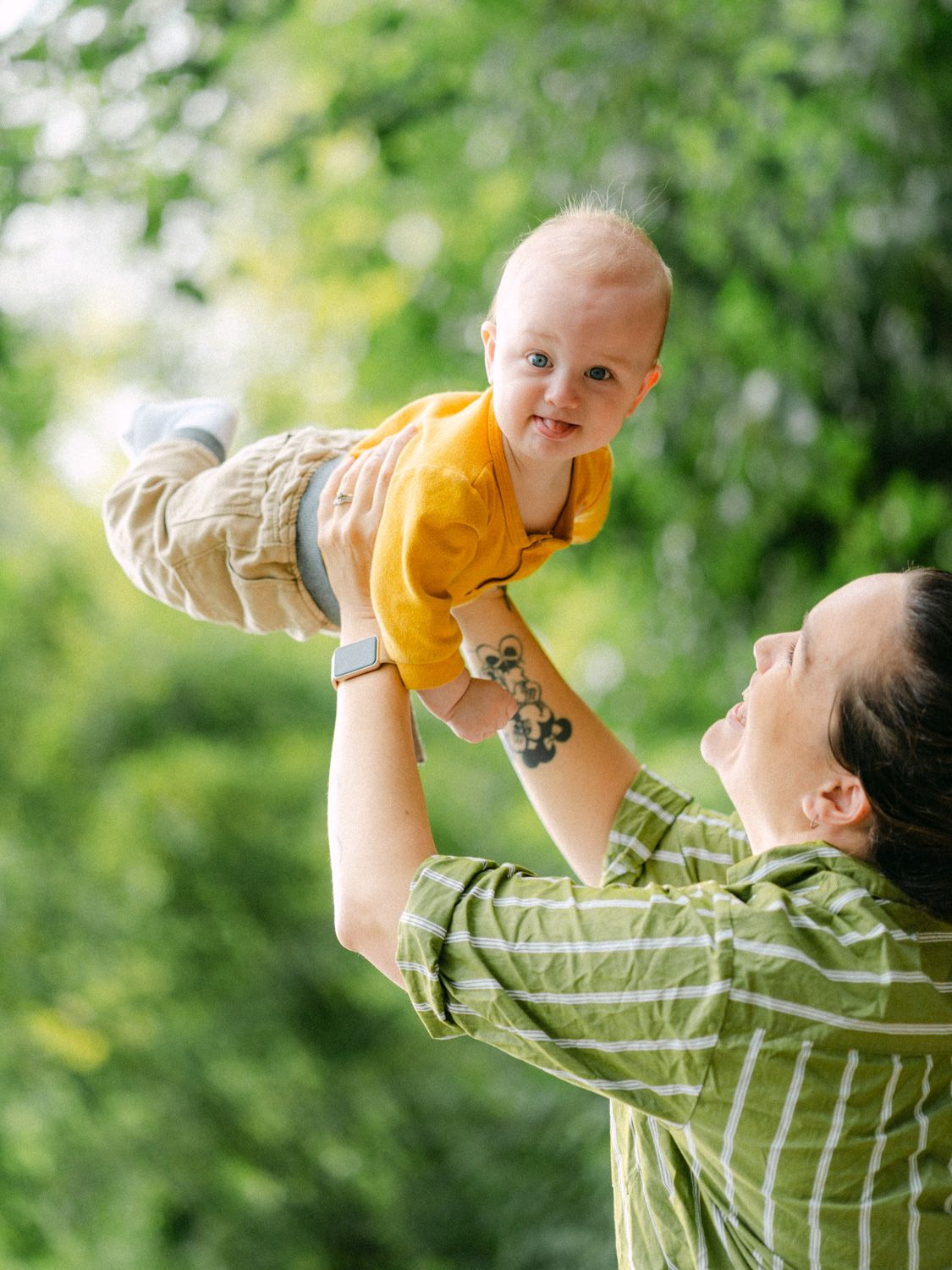 A smiling mother with a tattoo lifts her happy baby in a yellow onesie outdoors among lush green trees.