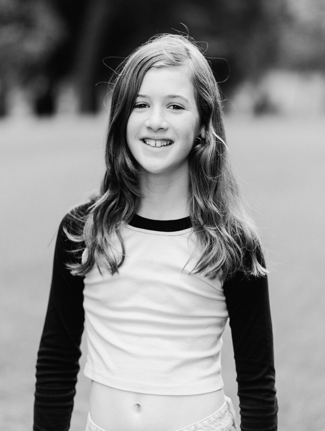 Black and white portrait of a smiling young girl with long hair wearing a two-tone shirt outdoors.