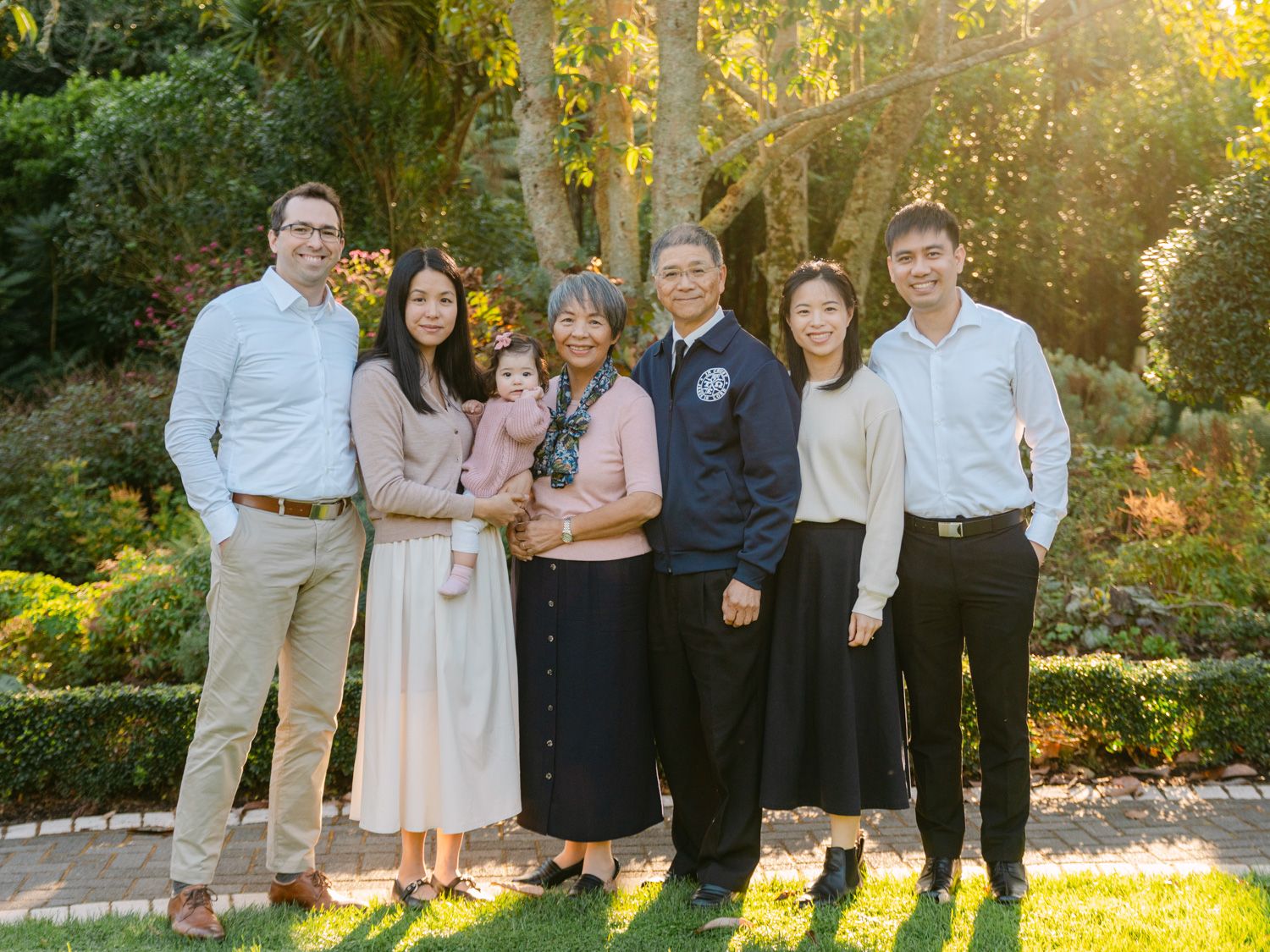 Multi-generational Asian family posing together outdoors in a sunlit garden, smiling for a portrait photo.
