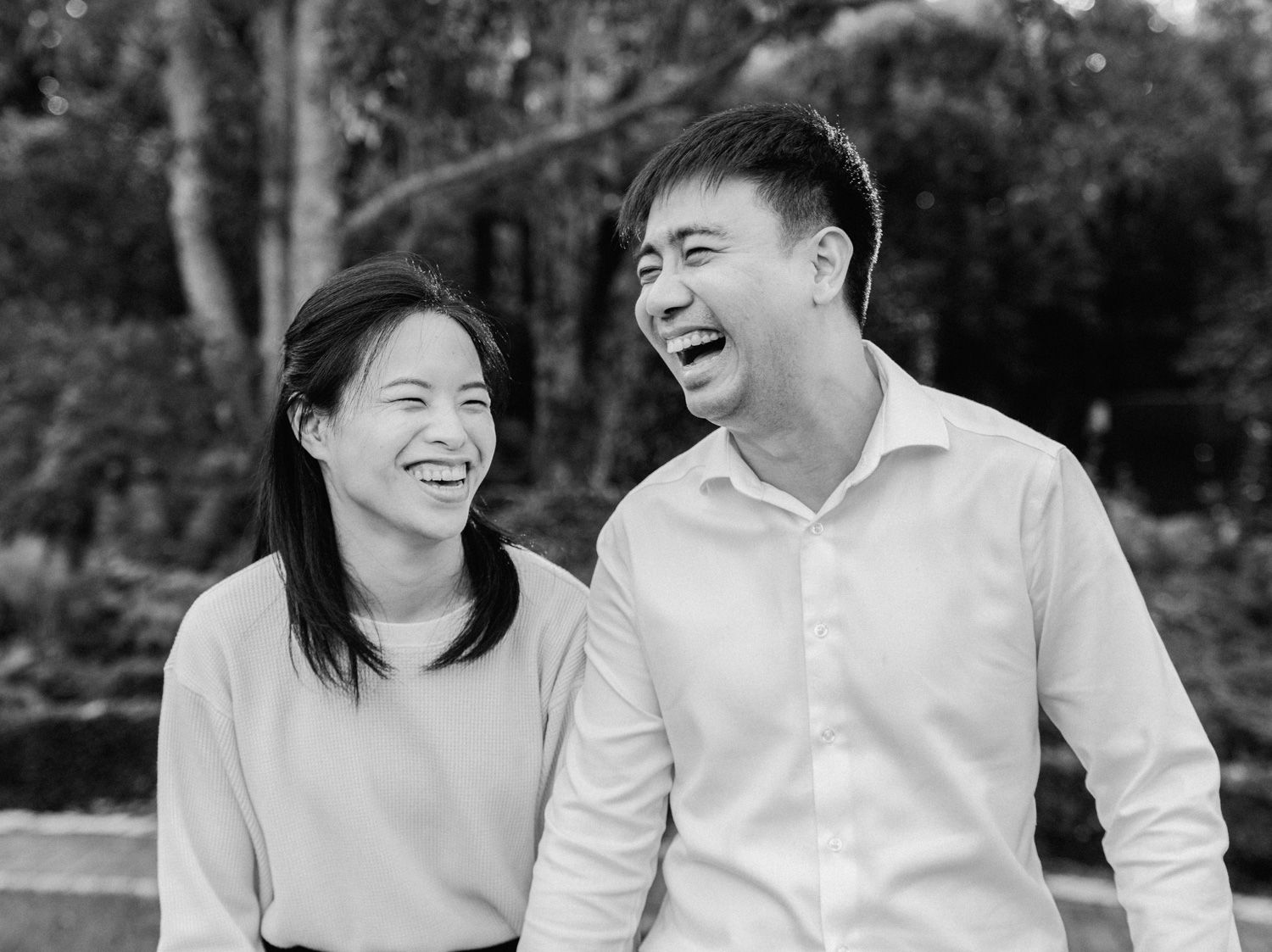 Black and white photo of a laughing Asian couple sharing a joyful moment outdoors among lush greenery.