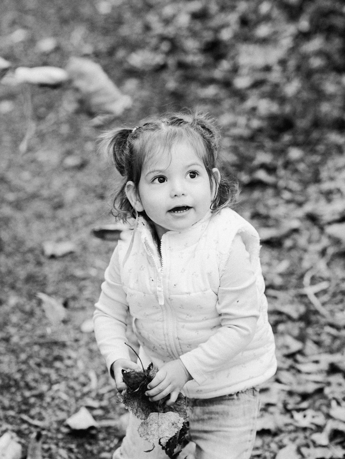 Black and white photo of a cute toddler girl with pigtails holding a leaf outdoors in a wooded area.