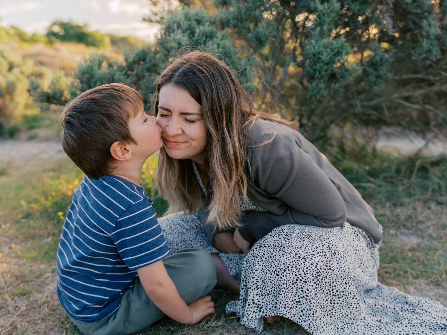 A young boy kisses his smiling mother on the cheek while sitting outdoors in a sunny green field.