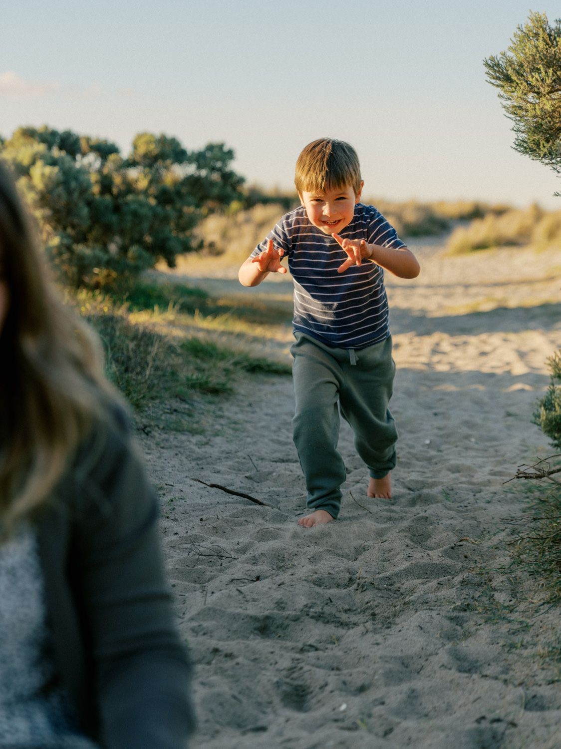 A barefoot boy in a striped shirt runs playfully on a sandy beach path toward the camera at golden hour.