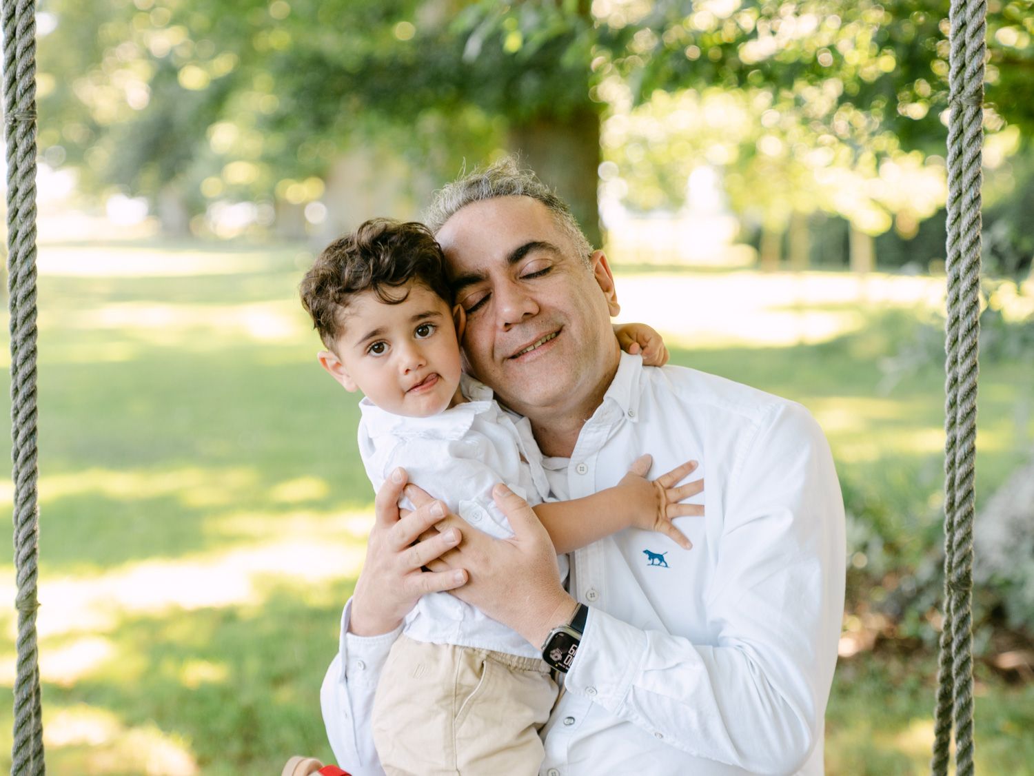 Man in white shirt hugging curly-haired toddler on a rope swing in a lush green park.