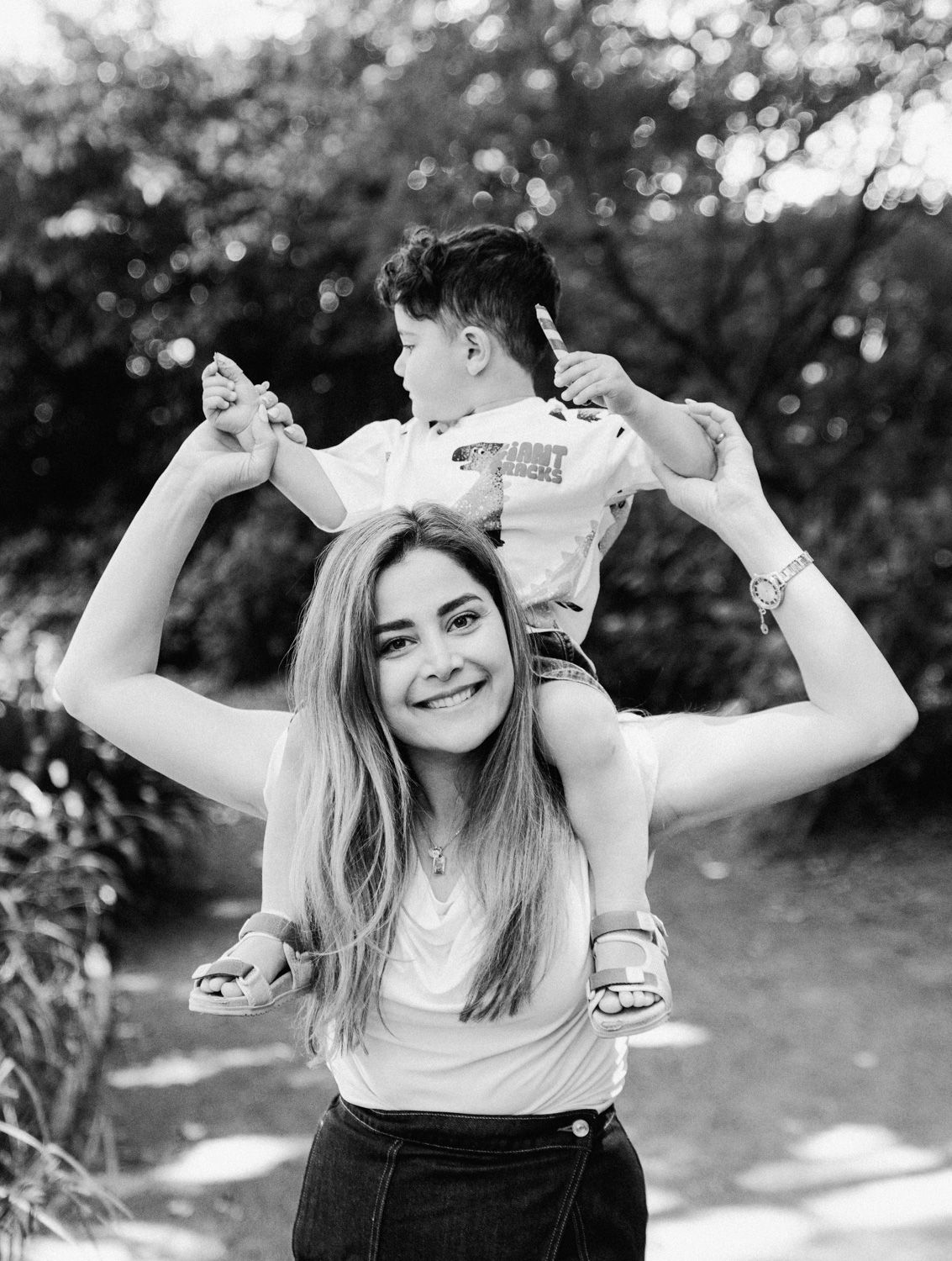 Black and white photo of a smiling young woman carrying a small boy on her shoulders outdoors.